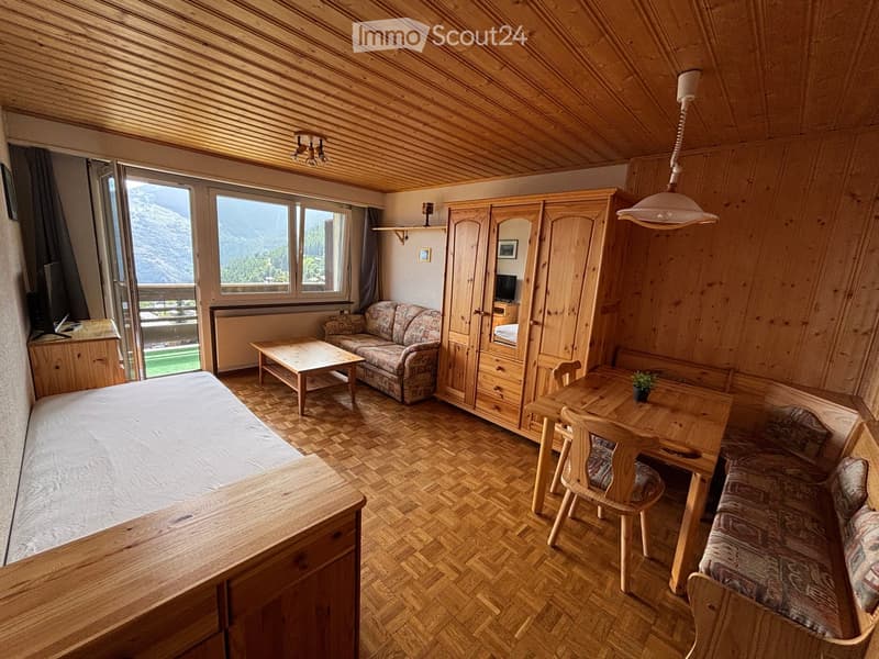 Cozy, wooden-furnished room with a bed, sofa, coffee table, dining table, and wardrobe, featuring large windows with a mountain view.