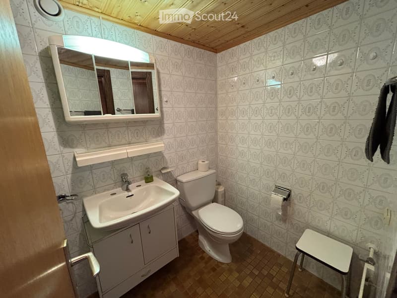 Bathroom with a sink, toilet, mirror, and a small stool. The walls are tiled with a patterned design, and the floor is tiled in a warm tone.