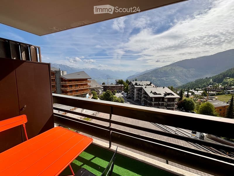 Balcony with a scenic mountain view, featuring an orange table and chair, and a small artificial grass area.