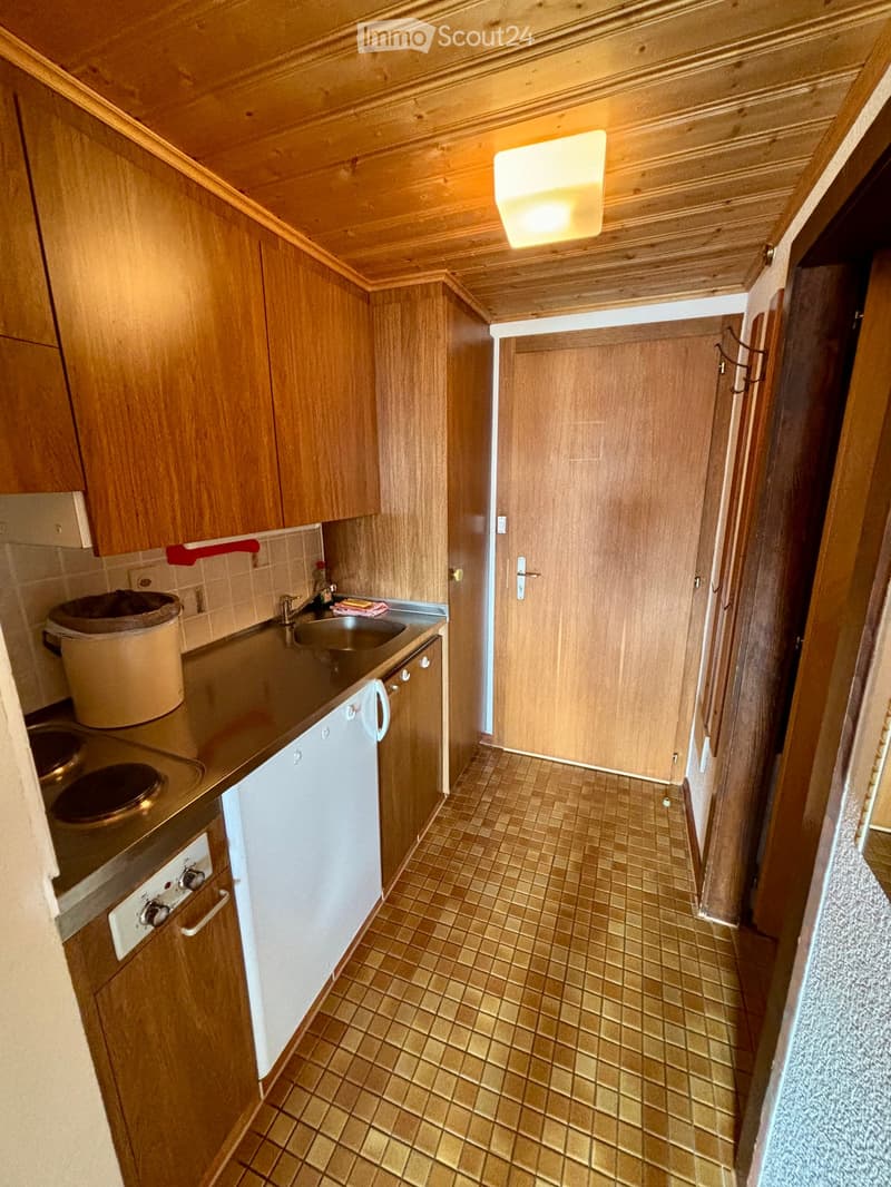 Compact kitchen with wooden cabinets, tiled floor, sink, stove, and refrigerator. Features a small countertop and built-in storage.