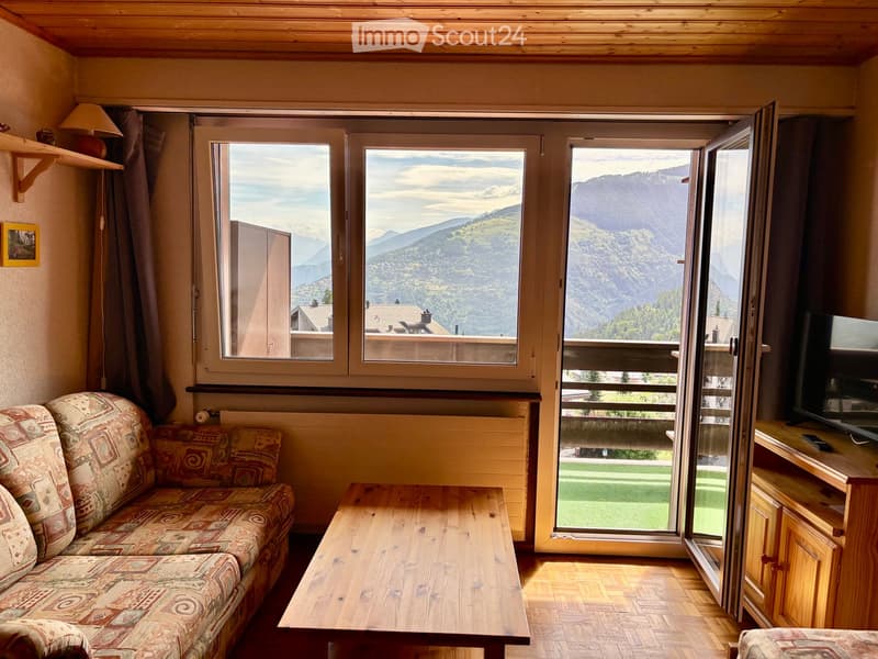 Cozy living room with wooden floors, large windows offering mountain views, sofa, coffee table, TV stand, and lamp.