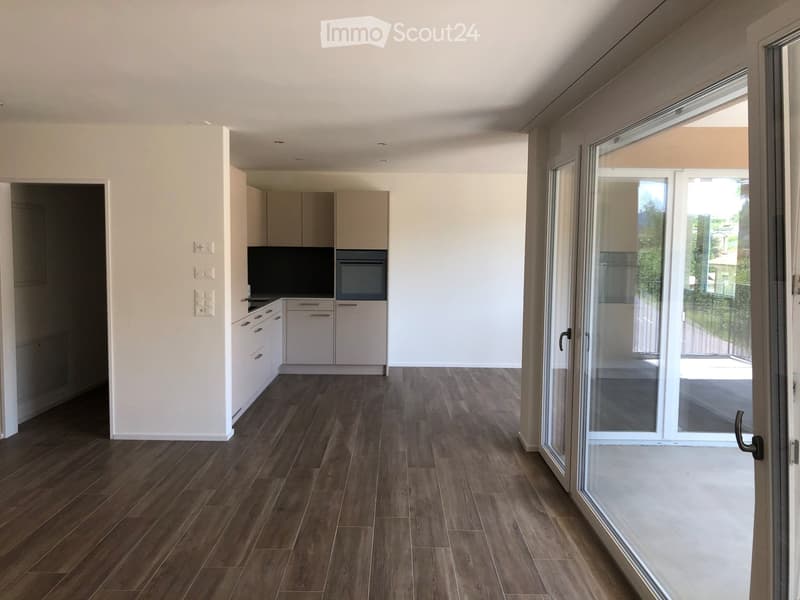 Empty room with wooden flooring, white walls, and a sliding glass door leading to a balcony. Kitchen area visible with cabinets and appliances.