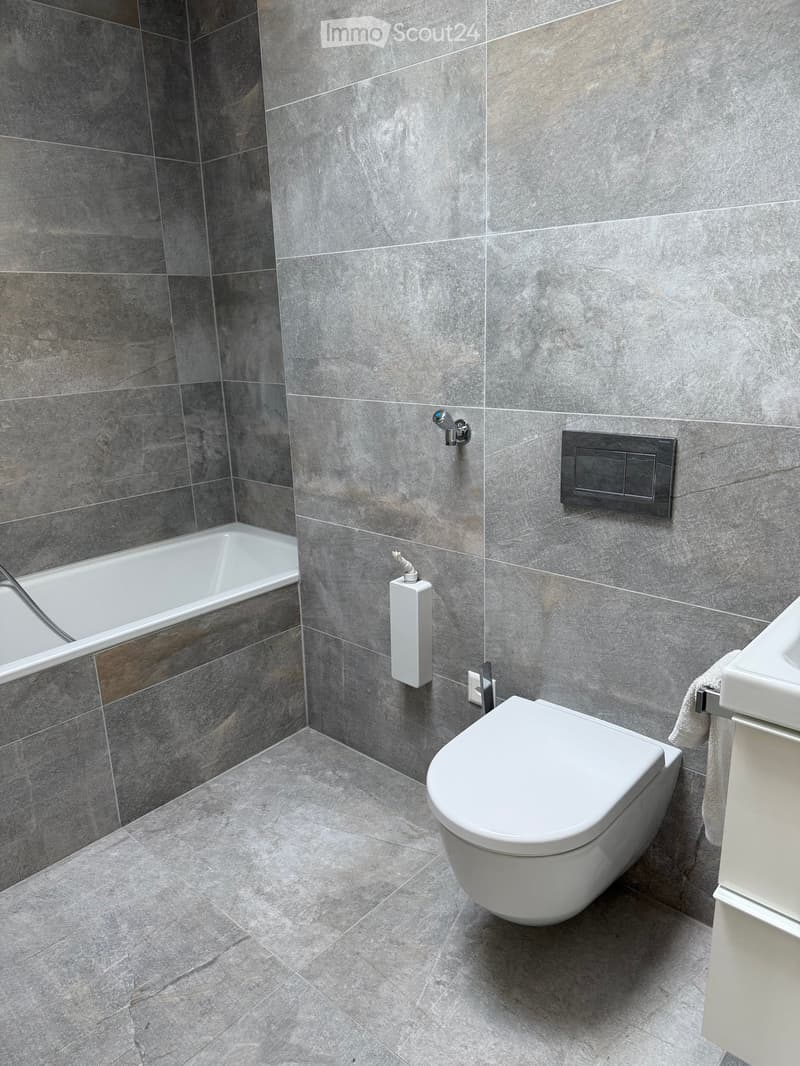 Modern bathroom with grey tiled walls and floor, featuring a toilet, bathtub, sink, and towel.