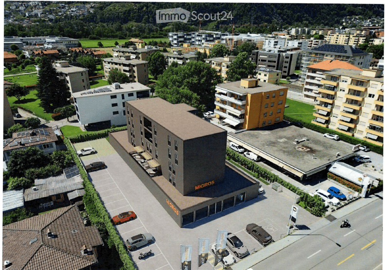Aerial view of a modern multi-story building labeled "MIGROS" with a large parking lot, surrounded by other buildings and greenery.