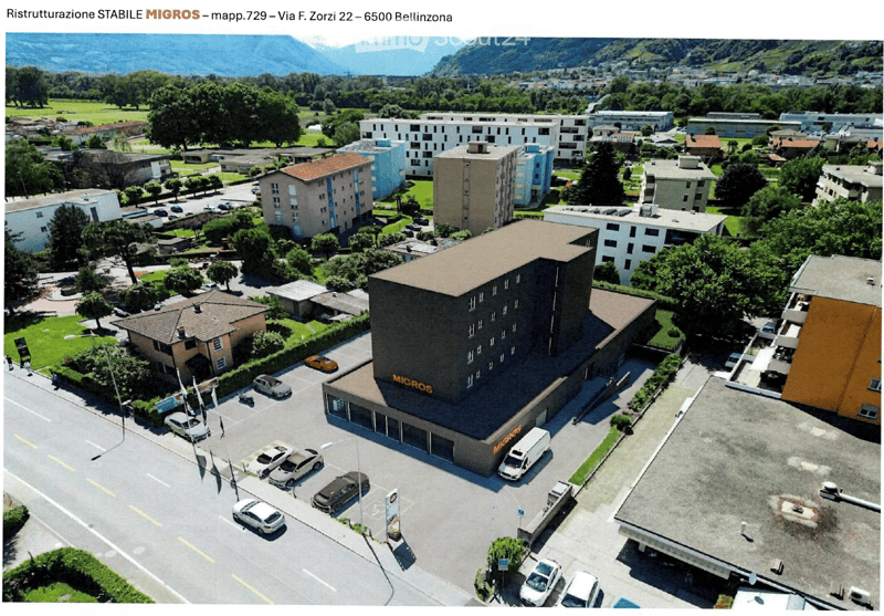 Aerial view of a multi-story building labeled "MIGROS" with a parking lot, surrounded by other buildings, greenery, and a mountainous backdrop.