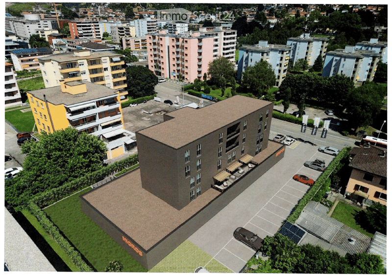 A modern multi-story building with a flat roof, surrounded by a parking lot and greenery, located in an urban area with other buildings and trees in the background.