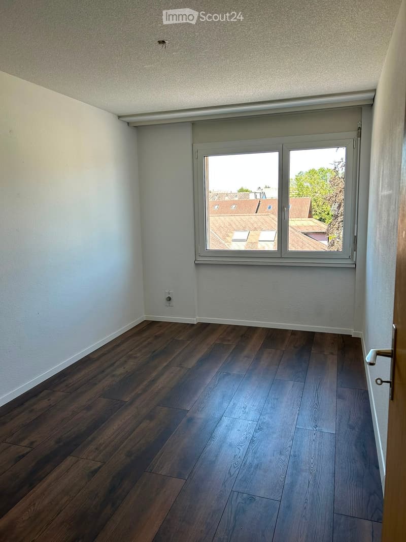 Empty room with wooden flooring, white walls, and a large window offering a view of rooftops and greenery.
