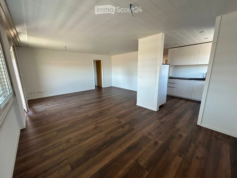 Empty room with wooden flooring, white walls, and a small kitchen area with white cabinets and a countertop.