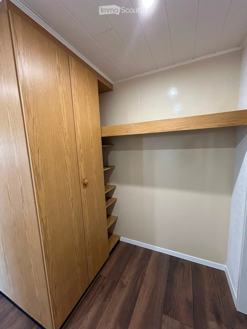Empty closet with wooden shelves and a closed wooden door, light-colored walls, and wooden flooring.