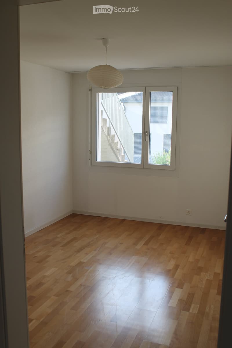 Empty room with wooden flooring, white walls, and a large window with a view of a staircase and outdoor area.