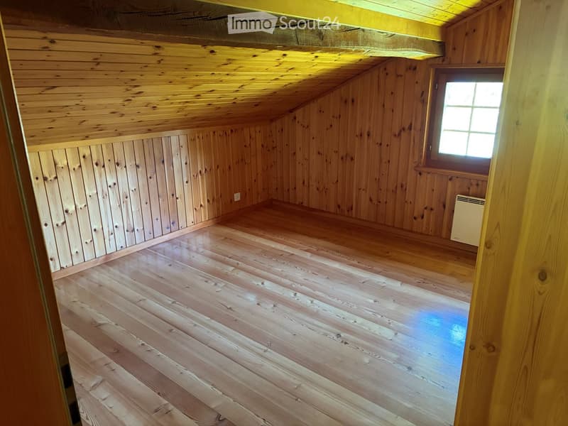 Empty wooden room with slanted ceiling, wooden walls, and a single window letting in natural light. The floor is made of polished wooden planks.