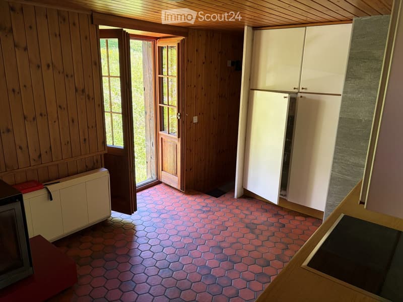 Bright kitchen with wooden walls, red hexagonal tiles, white cabinets, and a refrigerator. Features a large window and a door leading outside.