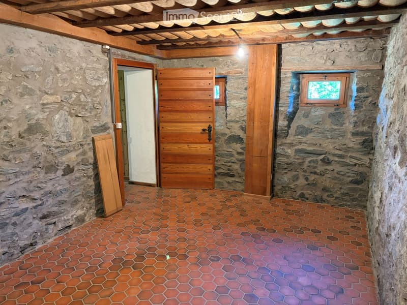A rustic stone-walled corridor with wooden beams, a wooden door, and a small window showing greenery outside.