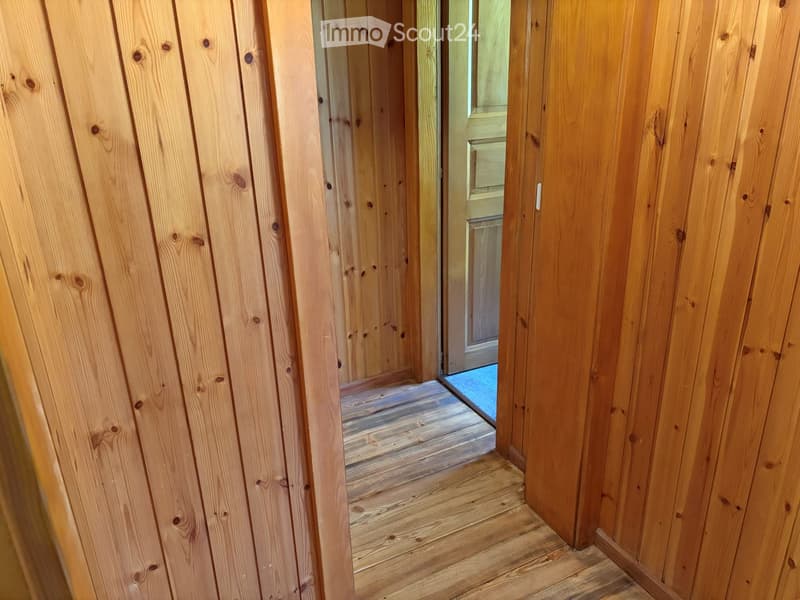 A narrow wooden hallway with vertical wooden panel walls and a wooden floor, leading to an open doorway.