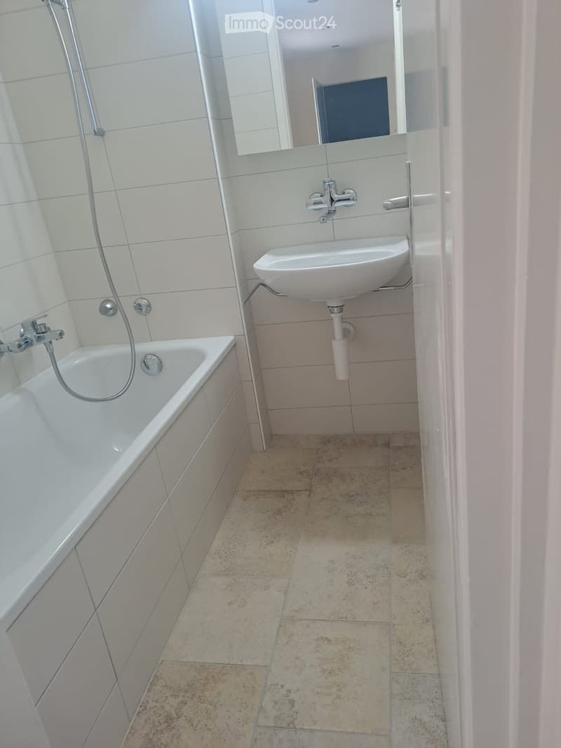 Modern bathroom with a white bathtub, a sink with a modern faucet, and a mirror above the sink. The floor is tiled with beige tiles.
