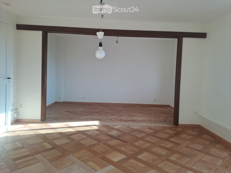 Empty room with wooden flooring and white walls, featuring exposed wooden beams and a hanging light fixture.