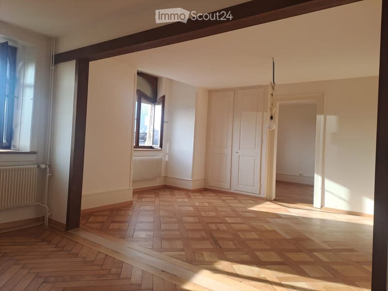 Empty room with wooden herringbone flooring, white walls, and large windows allowing natural light. Features include radiators, doors, and a hanging light fixture.