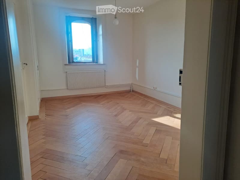 Empty room with wooden herringbone flooring, white walls, a window with a radiator below, and a hanging light fixture.
