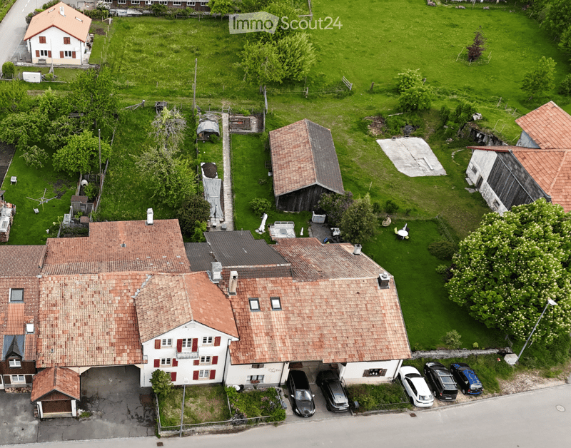 Aerial view of a two-story house with a red-tiled roof, multiple windows, and a garage. The property includes a well-maintained lawn, several outbuildings, and a parking area with several cars.