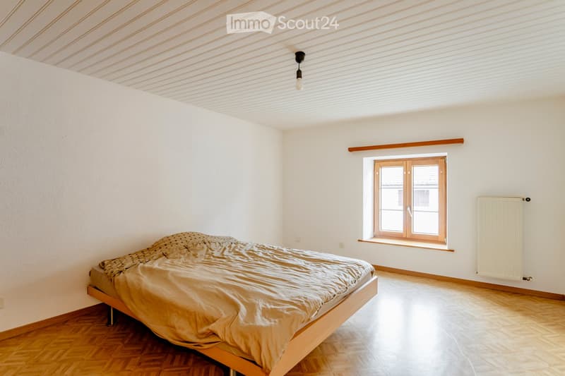 Bright bedroom with wooden flooring, white walls, a single bed with beige bedding, a window with wooden frame, and a radiator on the wall.