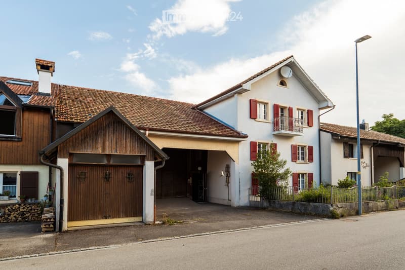 Two-story house with a tiled roof, white exterior, red shutters, and a wooden garage with a large wooden door.