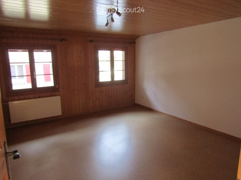 Empty room with wooden walls, carpeted floor, two windows, radiator, and a ceiling light fixture.