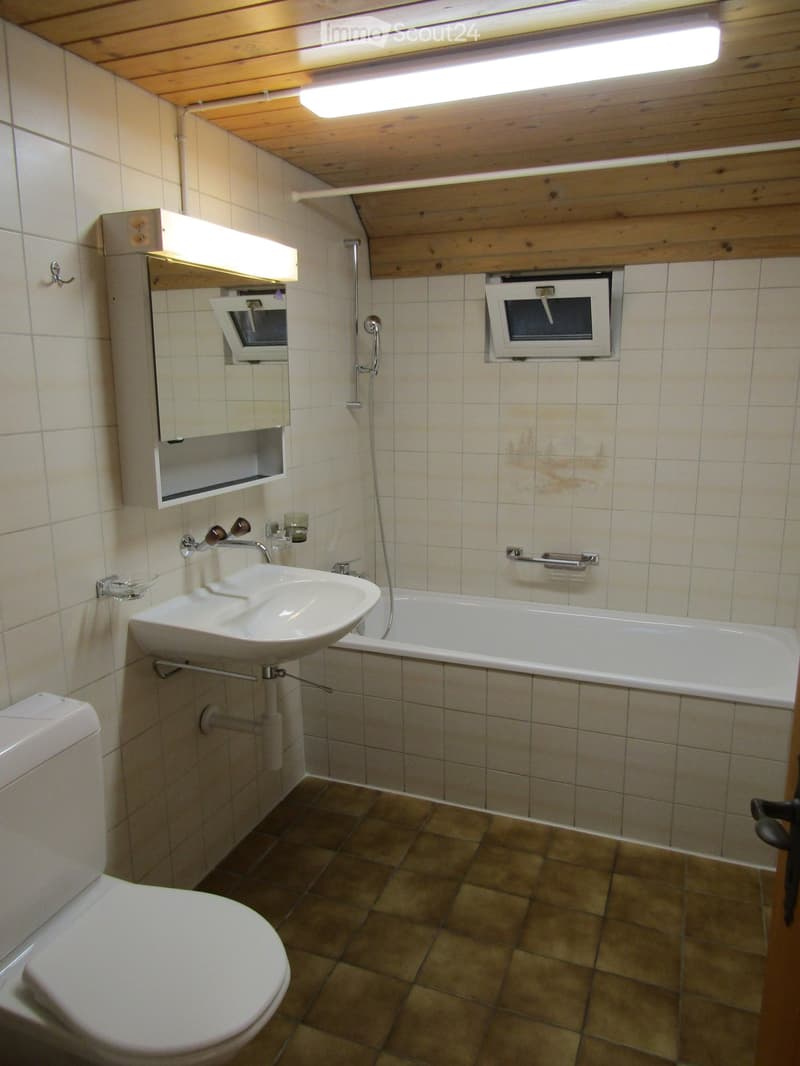 Bathroom with a toilet, sink, bathtub, and mirror. The walls are tiled, and the floor has brown tiles.
