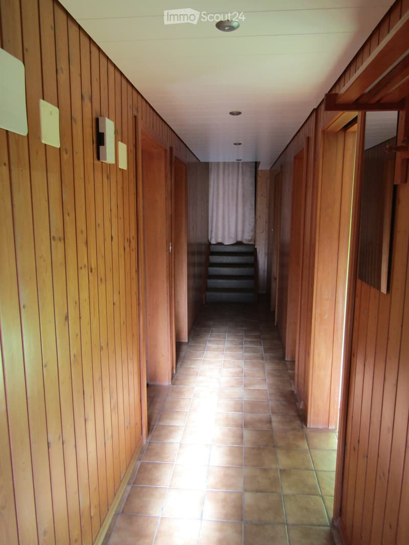 Wooden-paneled hallway with tiled floor, doors on both sides, and a staircase in the background.
