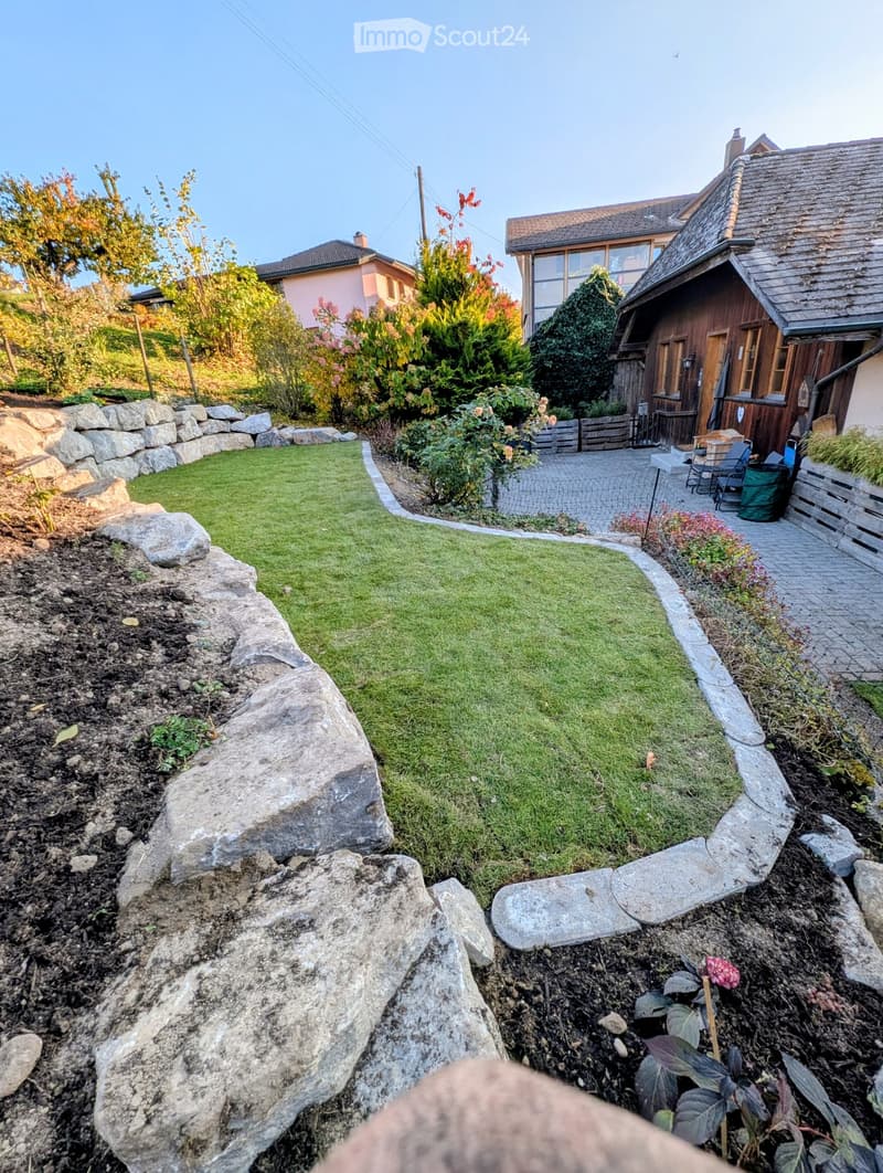 Well-maintained garden with a neatly trimmed lawn, stone border, large rocks, and a paved patio area with outdoor furniture near a wooden house.