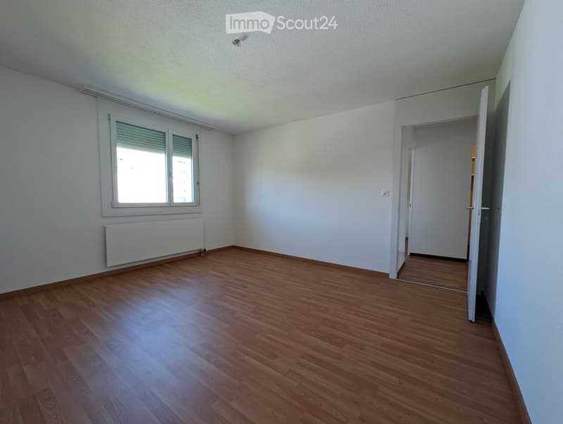 Empty room with wooden flooring, white walls, and a window with blinds. A radiator is visible below the window, and an open doorway leads to another space.