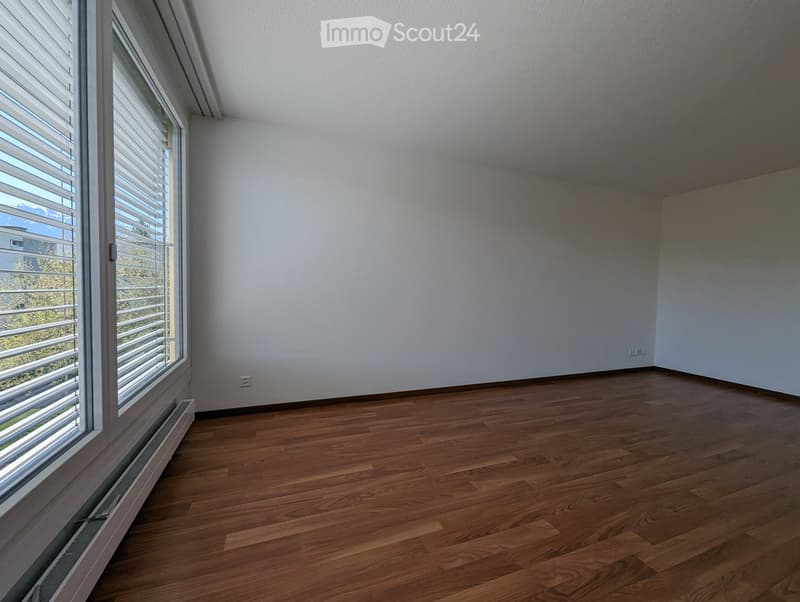 Empty room with wooden flooring, white walls, large window with blinds, radiator, and visible electrical outlets.