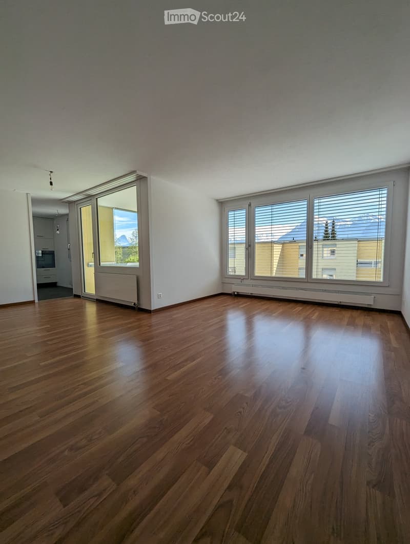 Empty room with wooden flooring, large windows with blinds, and a sliding glass door leading to another room.