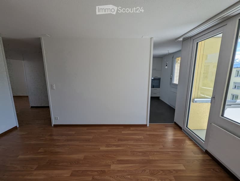 Empty room with wooden flooring, white walls, and large windows with blinds. The room is connected to a kitchen area visible in the background.