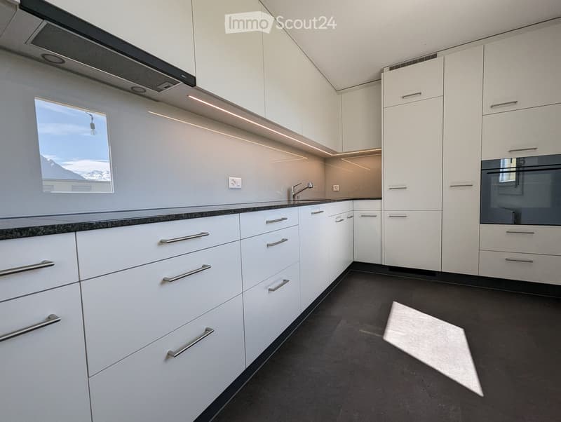 Modern kitchen with white cabinets, stainless steel handles, and a dark countertop. Features an integrated oven and a window with a scenic view.