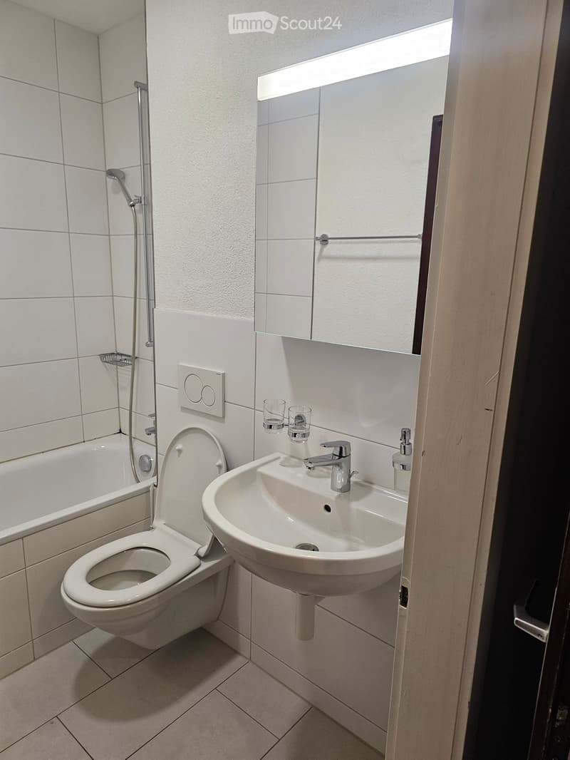 Modern bathroom with white tiles, a toilet, a sink with a modern faucet, a bathtub, and a showerhead. A large mirror with built-in lighting is mounted above the sink.