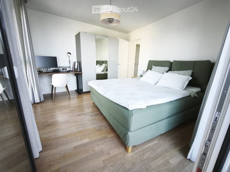Modern bedroom with a large bed, green sofa, desk with laptop, and a white chair. The room features light wooden flooring and white walls.
