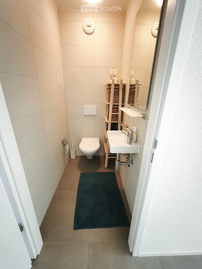 Modern bathroom with beige tiles, a toilet, a sink with a chrome faucet, a mirror, shelves with baskets, and a dark blue rug on the floor.