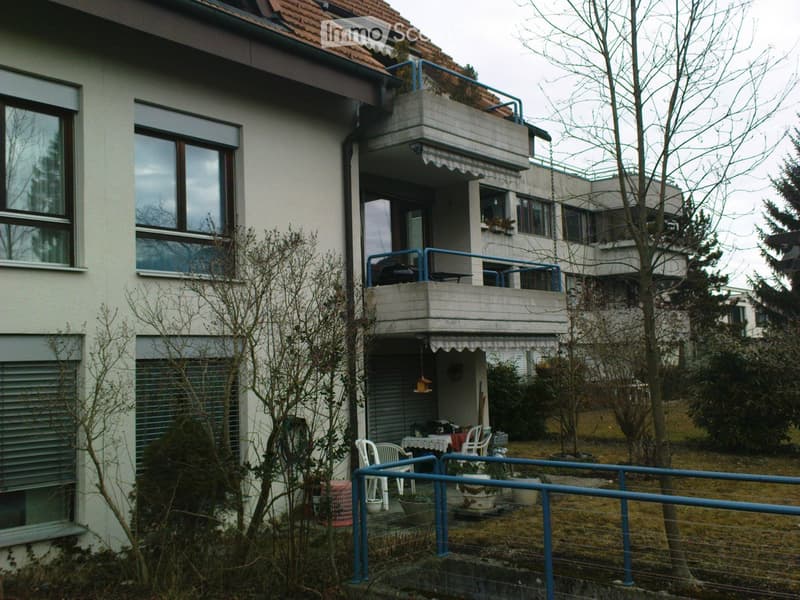 Two-story residential building with a balcony, large windows, and a small garden area with chairs and a table.