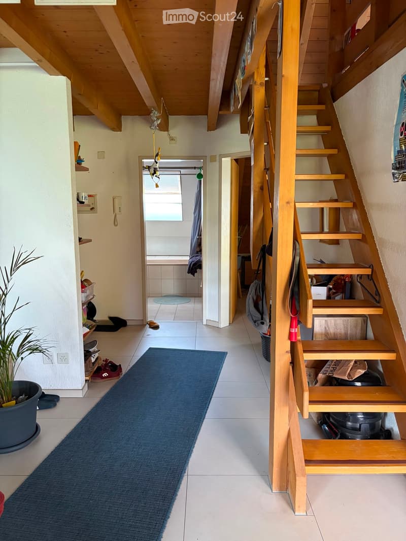 A narrow hallway with wooden stairs leading upwards, a blue runner rug, and a small plant in a pot. The hallway has white tiled flooring and walls, with a small shelf and some shoes visible on the left side.