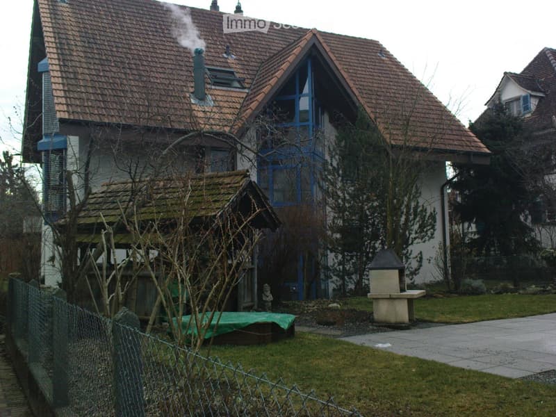 Two-story house with red-tiled roof, chimney, and blue-framed windows. The house has a small garden with a stone bench and a covered structure, surrounded by a fence.