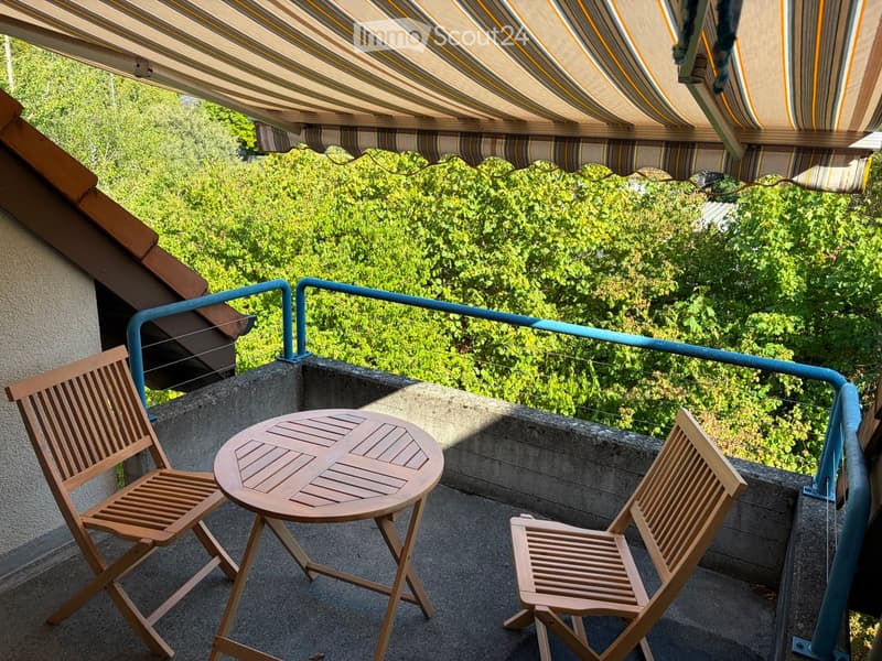Balcony with a round wooden table and two matching wooden chairs, under a striped awning, overlooking a green area.