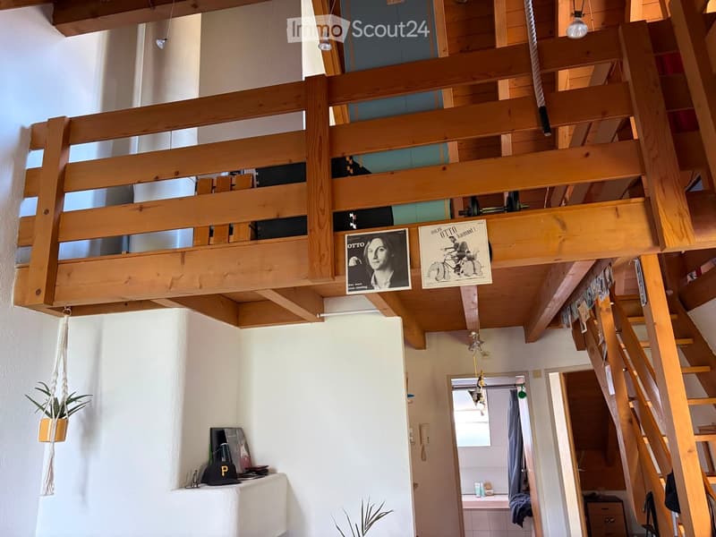 A modern, open-concept living space with exposed wooden beams and a lofted sleeping area. The lower level features a small table with a chair, a potted plant, and a poster on the wall. A staircase leads up to a lofted sleeping area with additional posters
