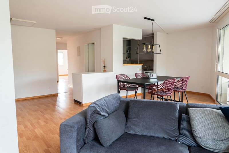 Living room with wooden floor, gray couch with pillows, dining table with four red chairs, kitchen area in the background, white walls, large windows