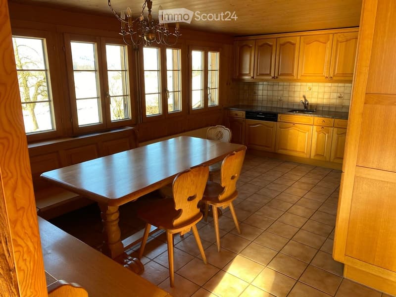 Bright kitchen with wooden cabinets, a large wooden dining table, and four matching chairs. The room features tiled flooring and large windows allowing natural light to fill the space.