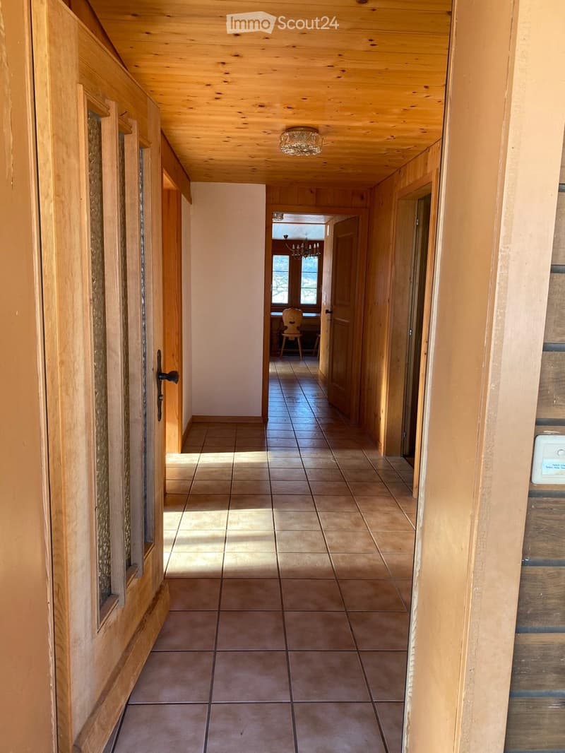 A long, narrow hallway with wooden walls and ceiling, tiled floor, and a chandelier hanging from the ceiling. The hallway leads to a room with a visible chair and table in the background.