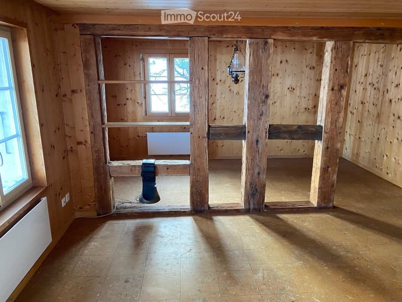 Empty room with wooden walls, exposed beams, and a tiled floor. Features include a large window, a radiator, and a hanging light fixture.