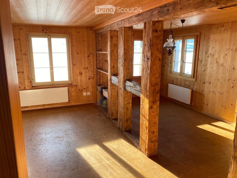 Empty room with wooden walls, exposed wooden beams, tiled floor, and large windows allowing natural light to enter. There are built-in wooden shelves and radiators on the walls.