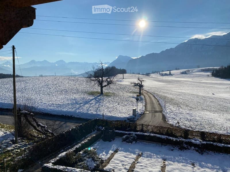 Snow-covered landscape with a road leading towards distant mountains, surrounded by trees and open fields.