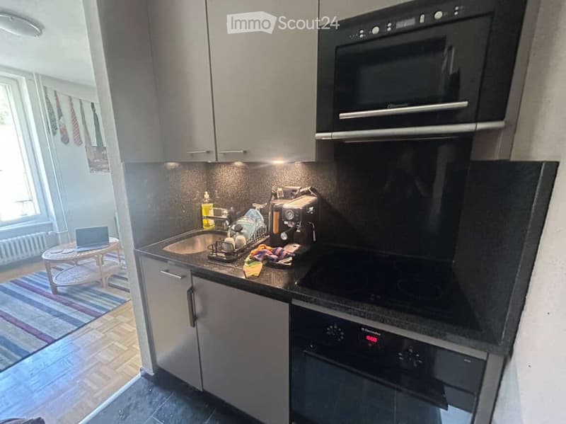 Modern kitchen with grey cabinets, black appliances, and a small round table with a laptop in the corner.