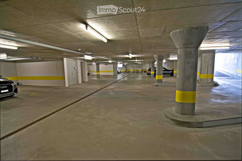 Underground parking area with concrete pillars, marked parking spaces, and several parked vehicles including cars and bicycles.